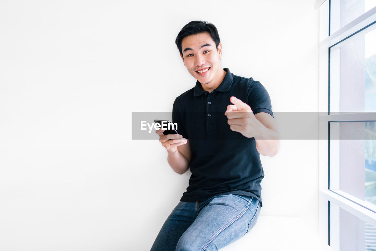 PORTRAIT OF YOUNG MAN USING PHONE WHILE STANDING ON WALL