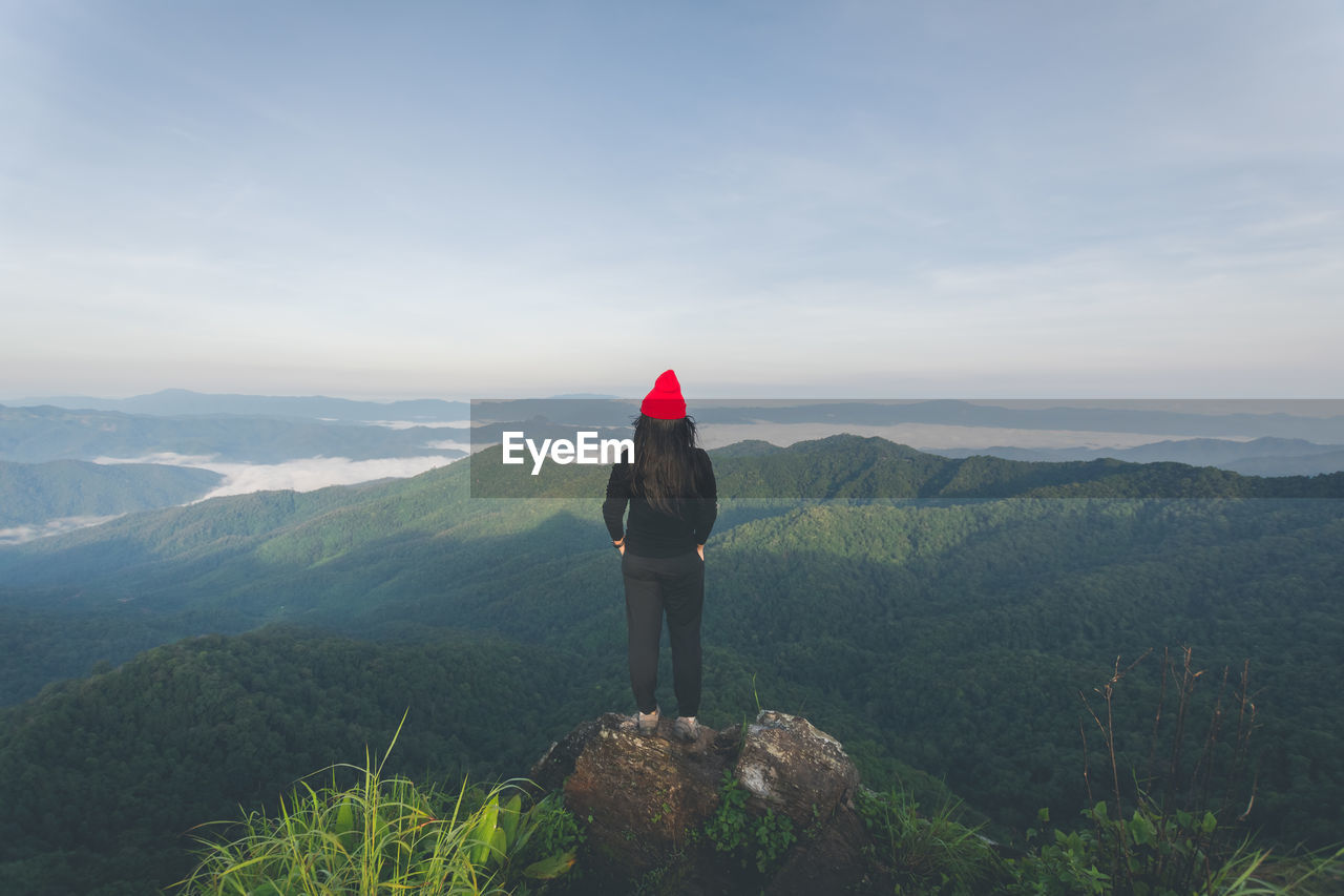 REAR VIEW OF WOMAN STANDING ON MOUNTAIN AGAINST SKY