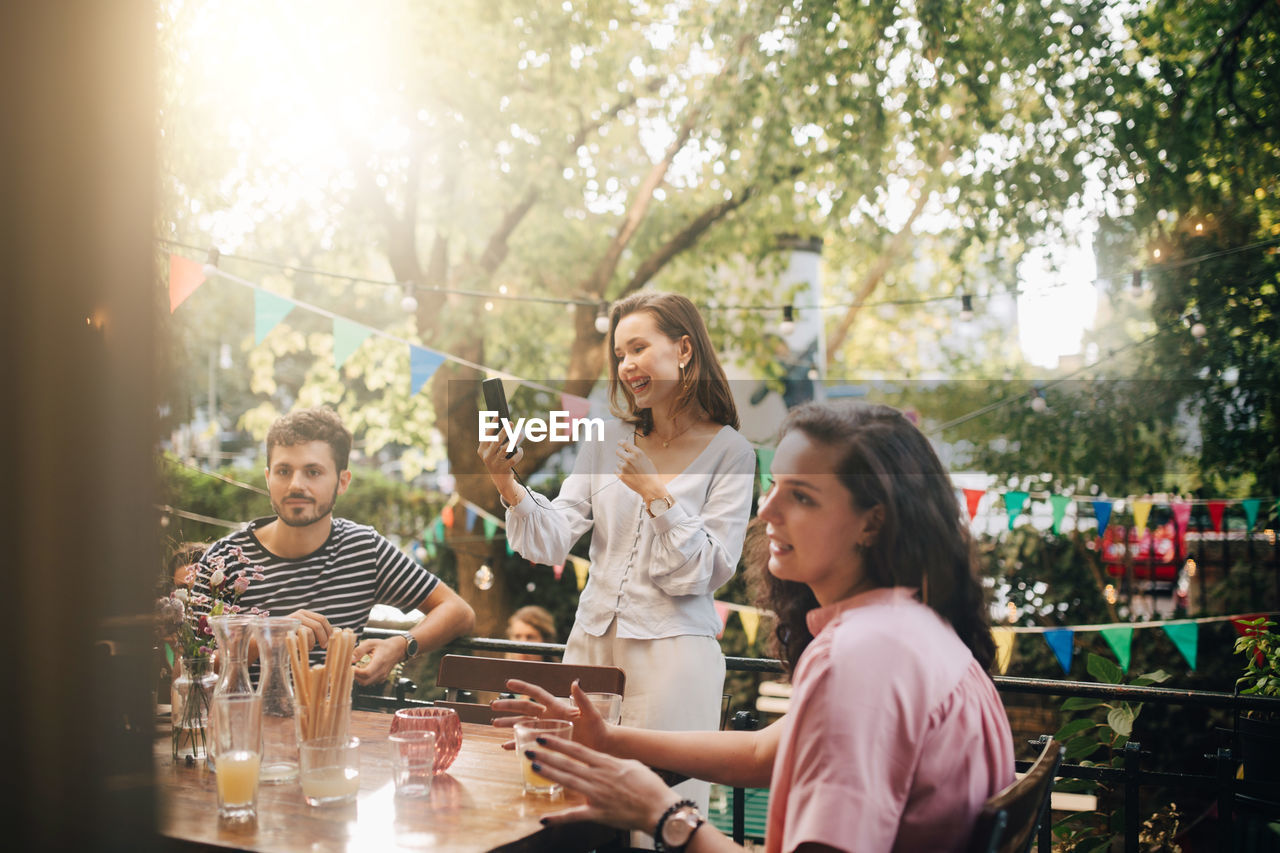 Smiling young woman video conferencing through smart phone while friends sitting in balcony during party