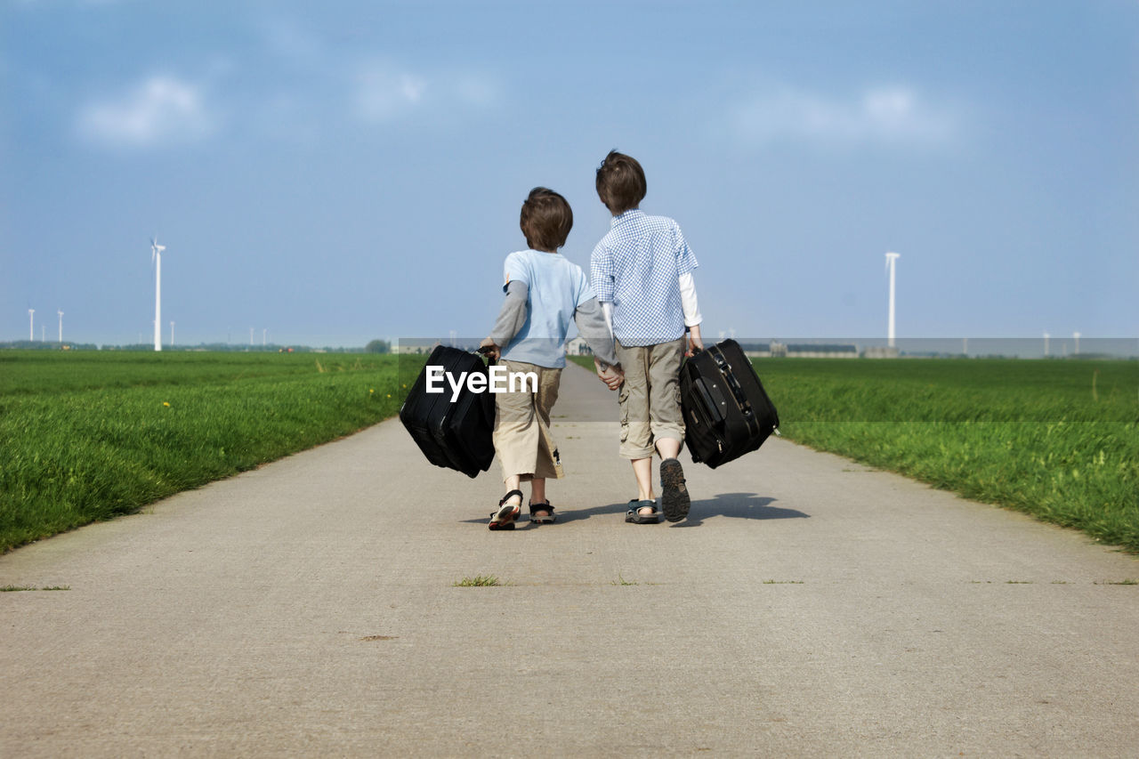 Rear view of brothers carrying suitcases while walking on road against sky