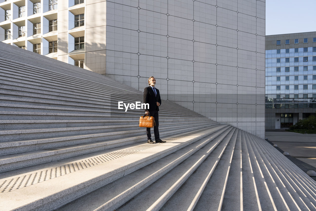 Mature businessman holding bag standing on staircase in front of building