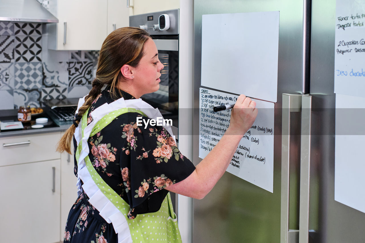 Side view of female in apron standing and writing notes on paper hanging on fridge while cooking in light kitchen in daytime