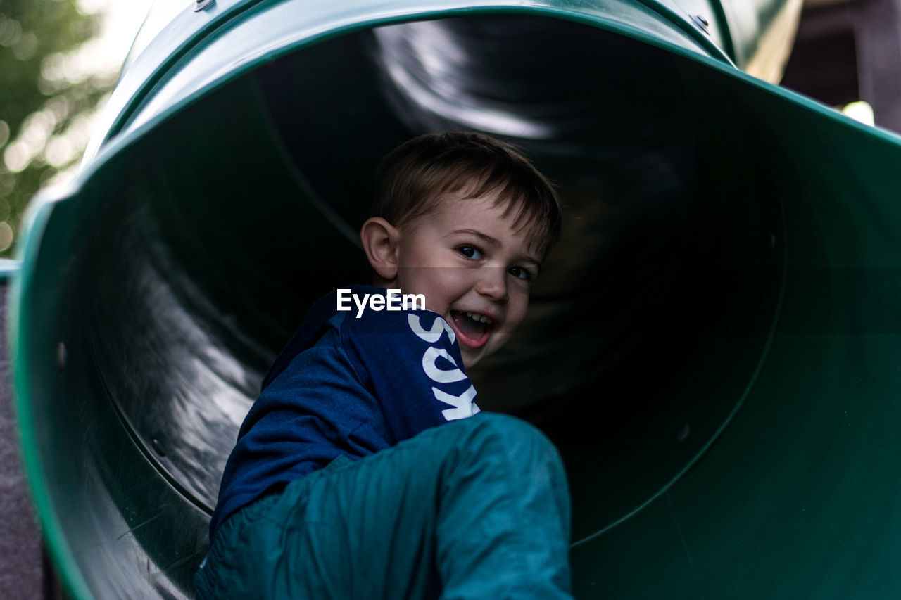Low angle portrait of cheerful boy sitting in slide