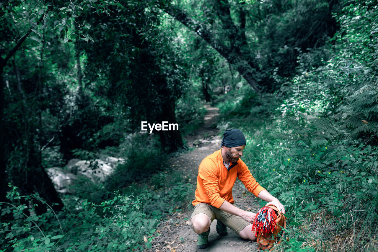 Calm bearded male tourist with backpack sitting on trail in green woods during summer vacation