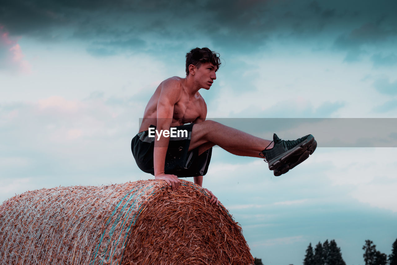 Low angle view of shirtless teenage boy exercising on hay bale against sky