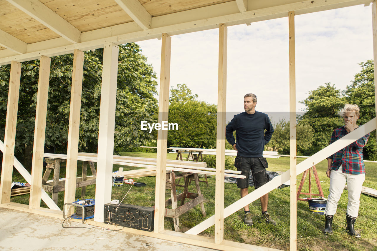 Serious couple looking at incomplete shed against trees