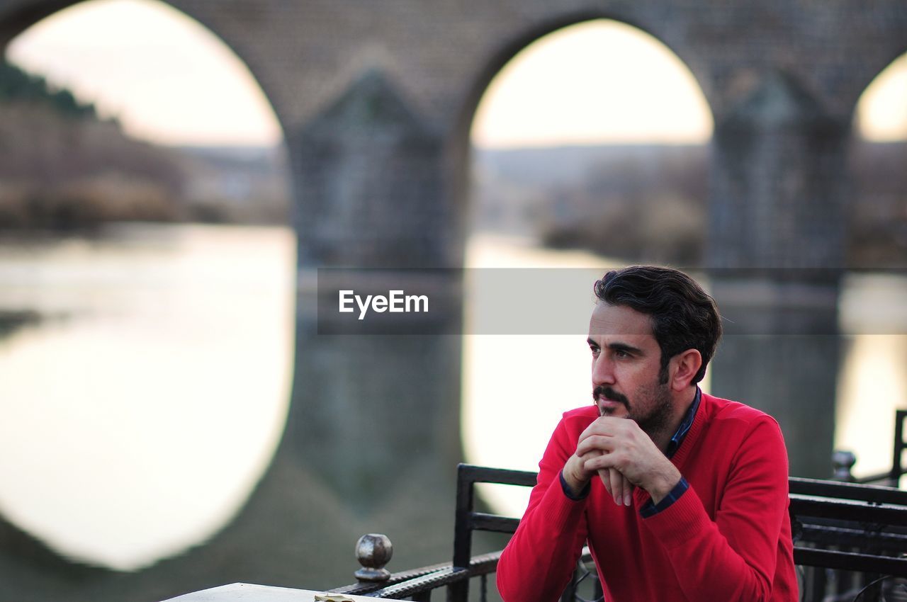 Thoughtful man looking away while sitting against river and bridge in city