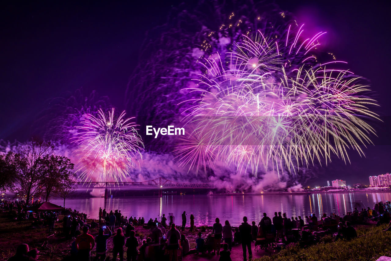 People looking at illuminated firework display over river at night