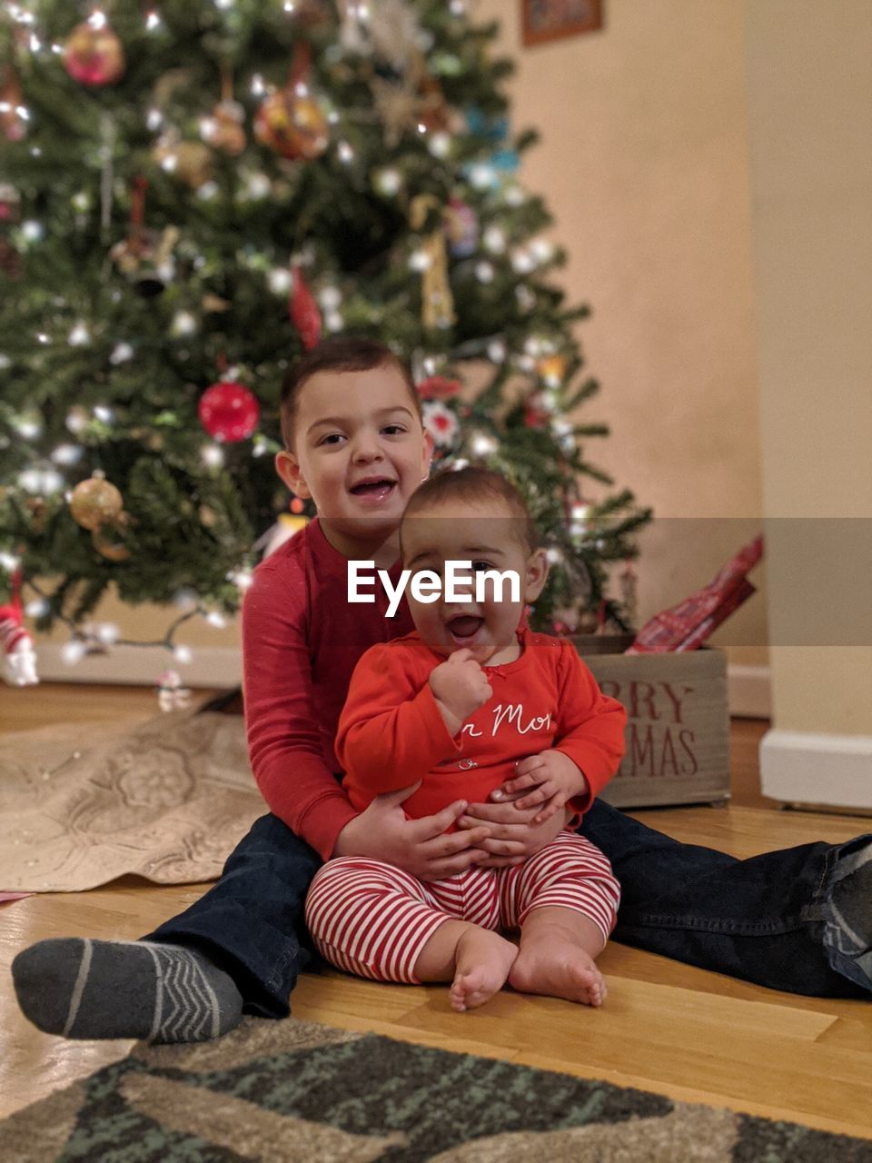 Full length portrait of smiling siblings sitting by christmas tree at home