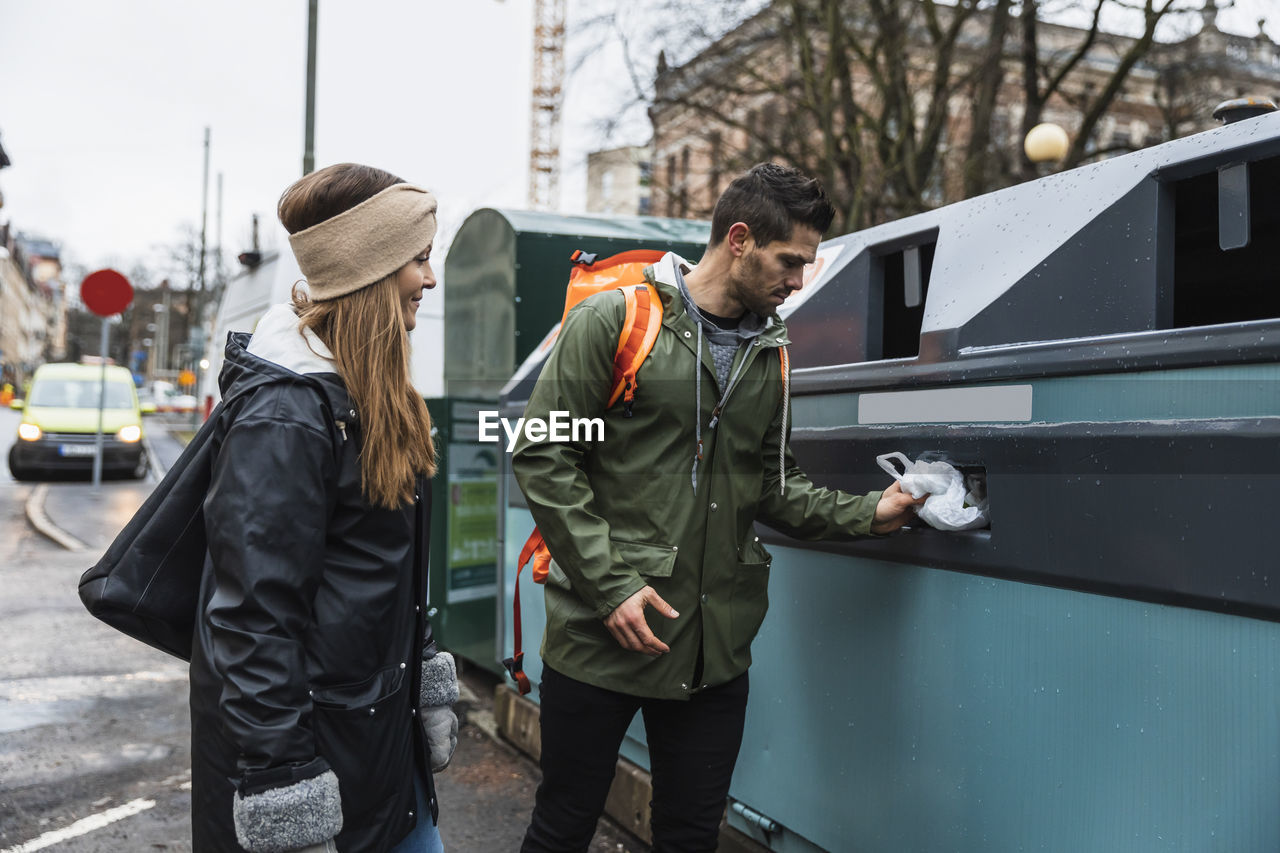 Heterosexual couple throwing plastic in recycling bin while standing at city