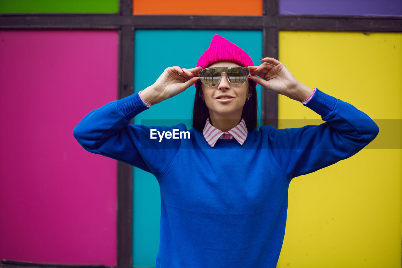 Fashionable brunette woman in a pink warm hat and sweater and sunglasses stands against background