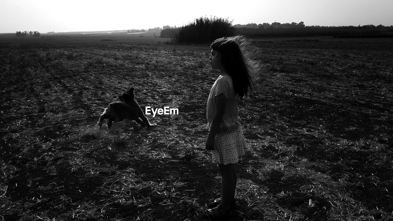 Side view of girl standing by dog running on land against sky