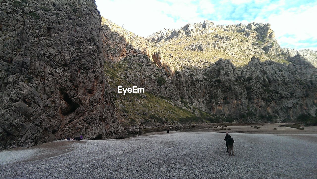 MAN WALKING ON ROAD AMIDST ROCKS AGAINST SKY