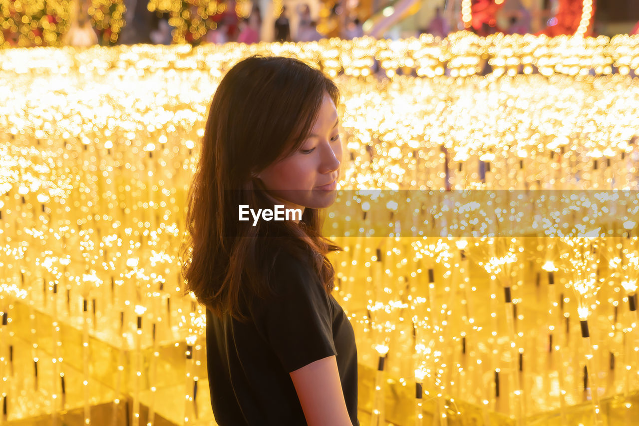 Mid adult woman looking down while standing against illuminated lighting equipment