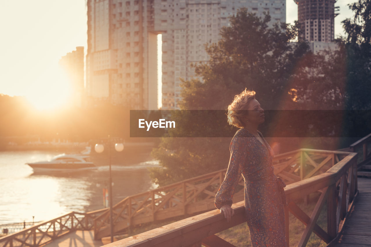 Woman leaning against railing while standing in city during sunset