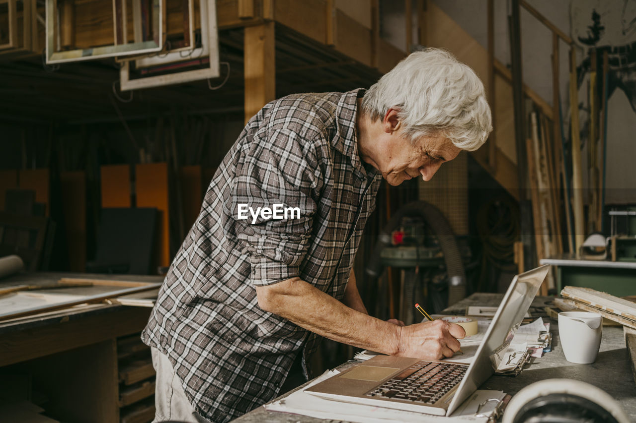 Senior male carpenter preparing document with laptop on workbench at repair shop