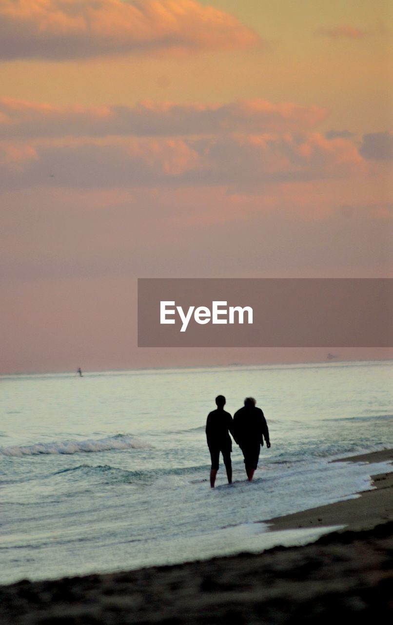 Silhouette couple walking on shore at beach against sky during sunset