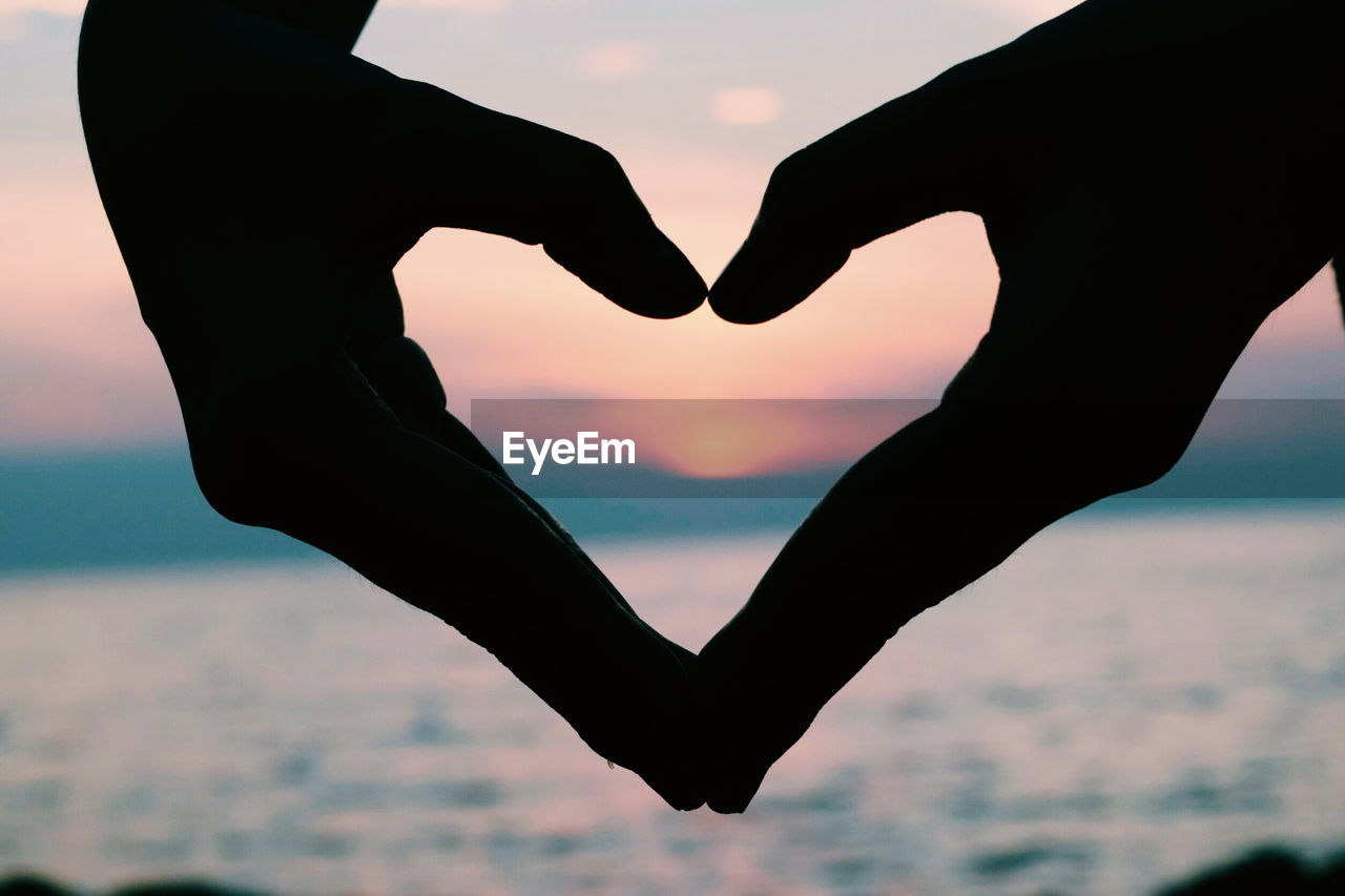 Silhouette hand making heart shape against sea during sunset