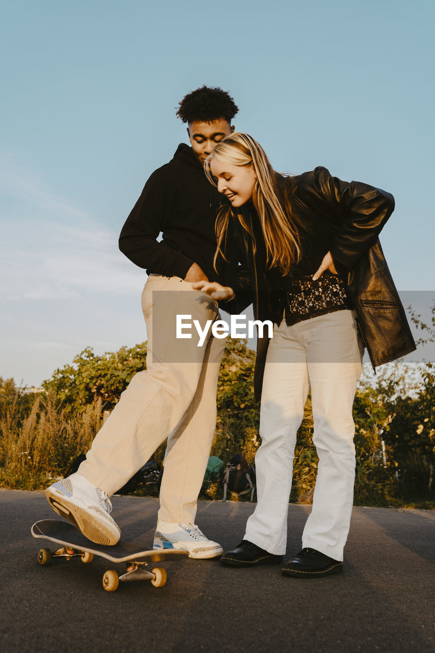 Male and female friends standing by skateboard in park during sunset