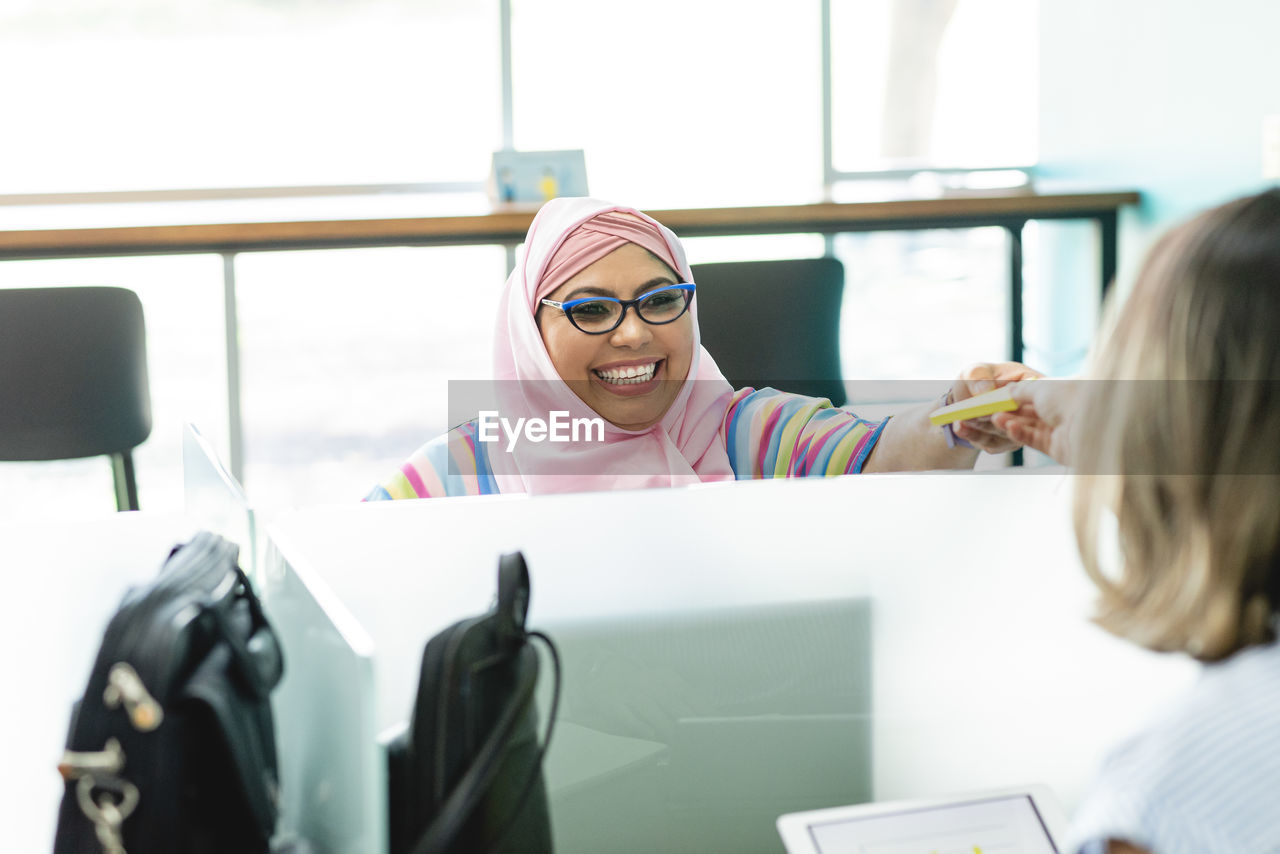 Positive muslim female in hijab sitting at table and giving paper stickers to unrecognizable coworker during work in office in costa rica in daytime