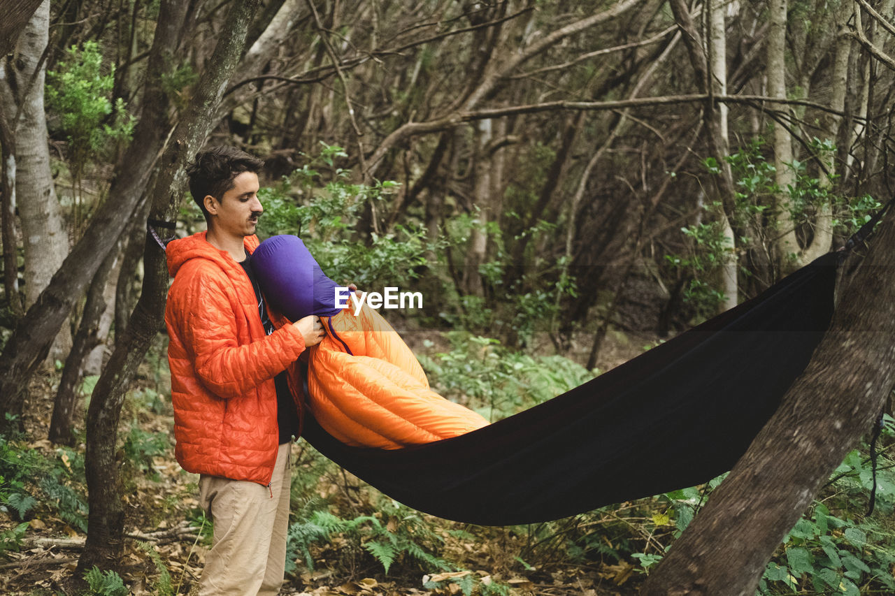 A man packs a sleeping bag and hammock at campsite in the forest