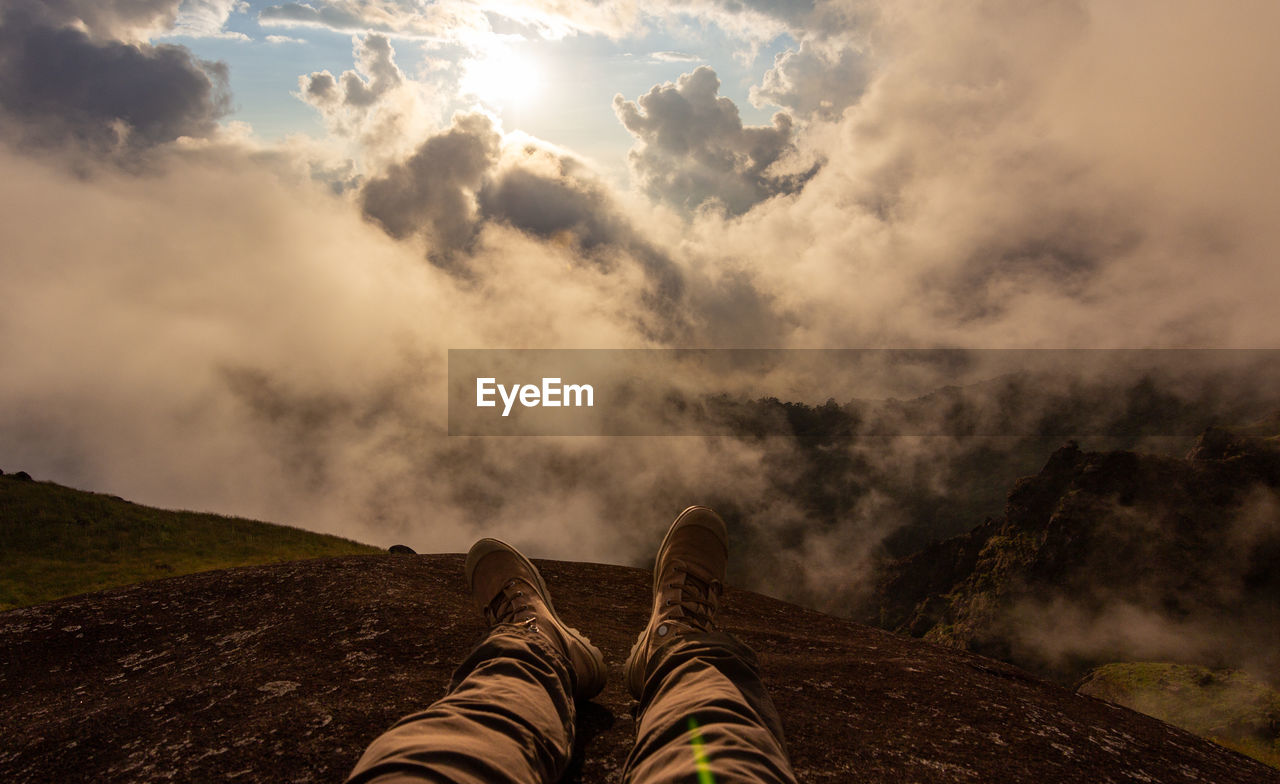 Low section of person sitting on mountain against cloudy sky during sunset