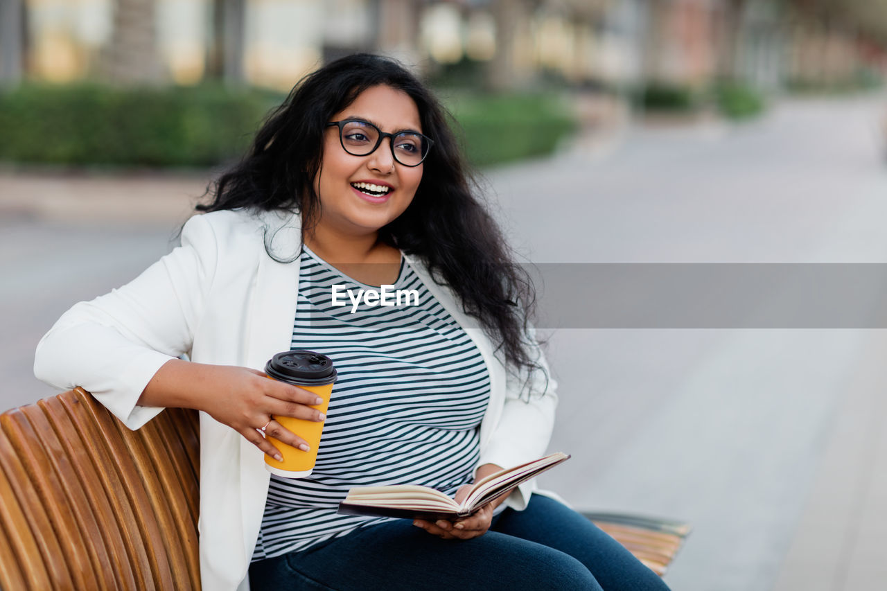 An indian girl is sitting on a bench, reading a book and drinking coffee.