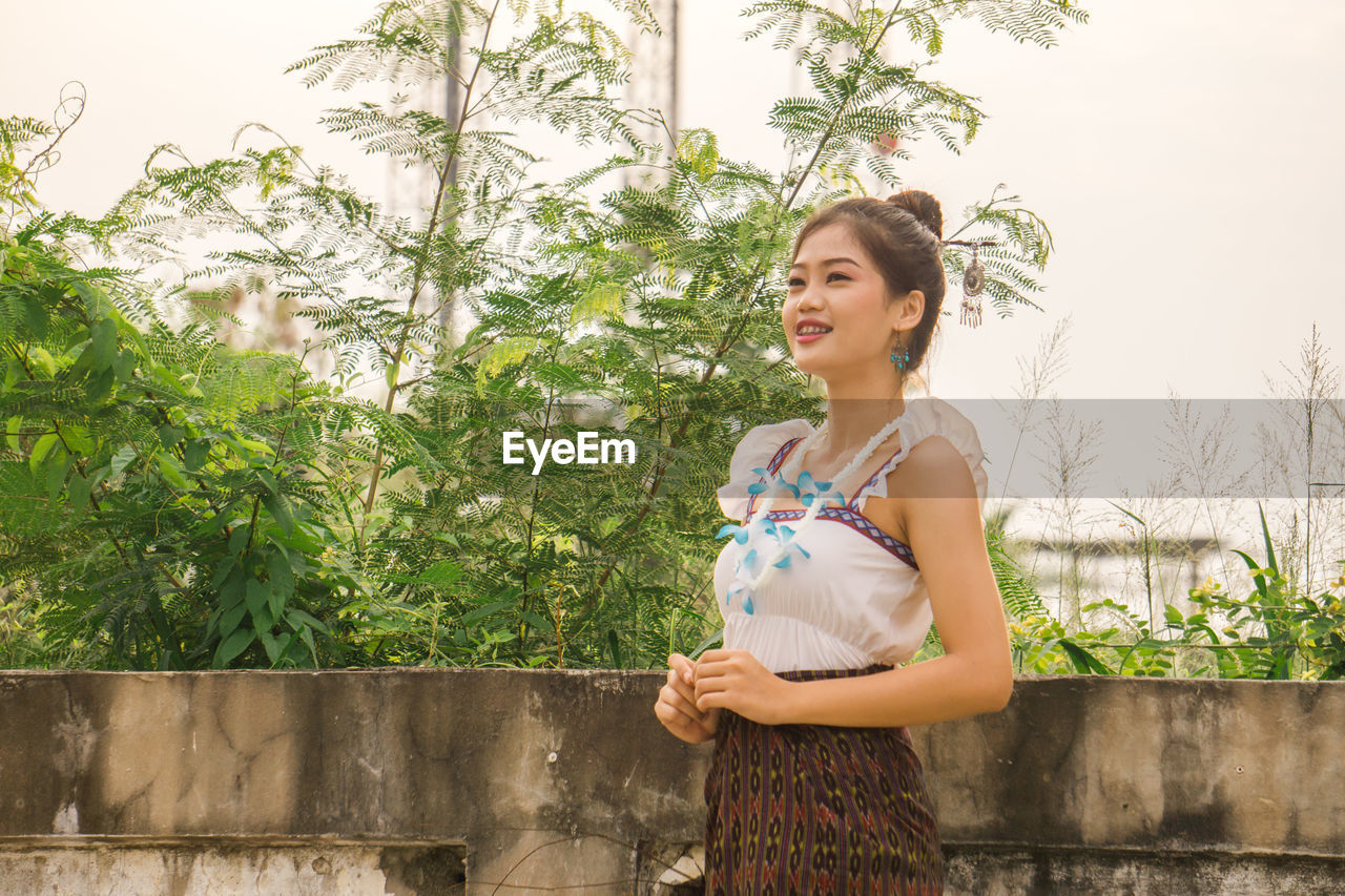 SMILING YOUNG WOMAN STANDING AGAINST PLANTS