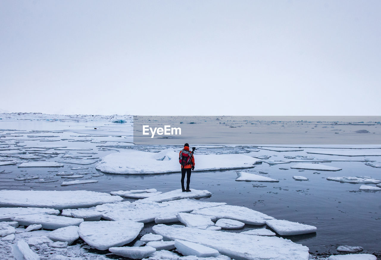 Back view of anonymous explorer in warm clothes standing on piece of ice in sea near shore and admiring winter seascape in iceland