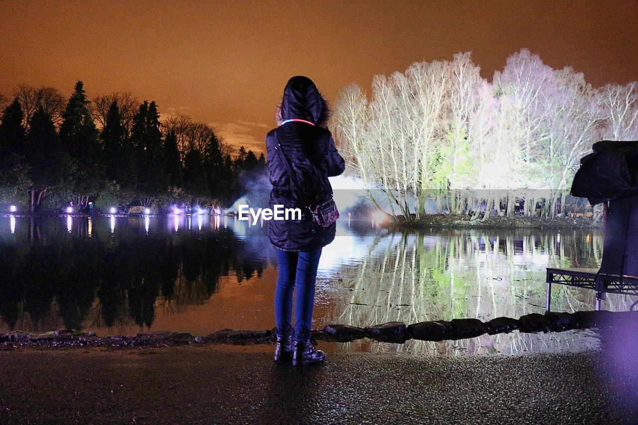 Rear view of woman standing by pond at rouken glen during night