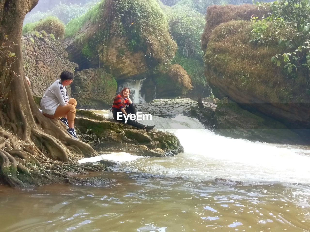 PEOPLE SITTING ON ROCKS BY RIVER