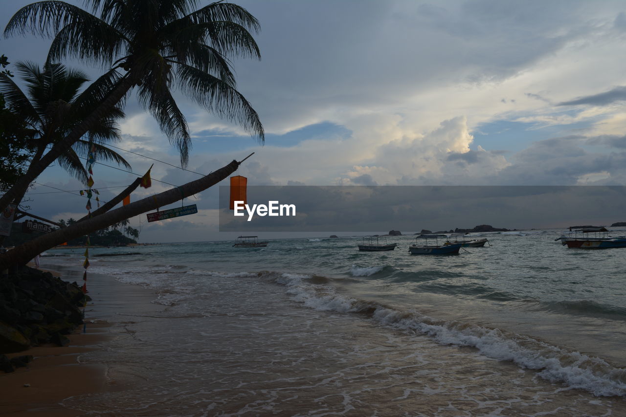 water, sea, sky, beach, palm tree, land, ocean, shore, tropical climate, cloud, coast, tree, nature, wave, body of water, scenics - nature, beauty in nature, sand, tranquility, coconut palm tree, nautical vessel, plant, tranquil scene, sunset, travel destinations, transportation, bay, travel, no people, outdoors, horizon over water, holiday, environment, idyllic, horizon, tropics, vacation, tropical tree, coastline, seascape, trip, dusk, tourism, mode of transportation, motion, wind wave, ship, island