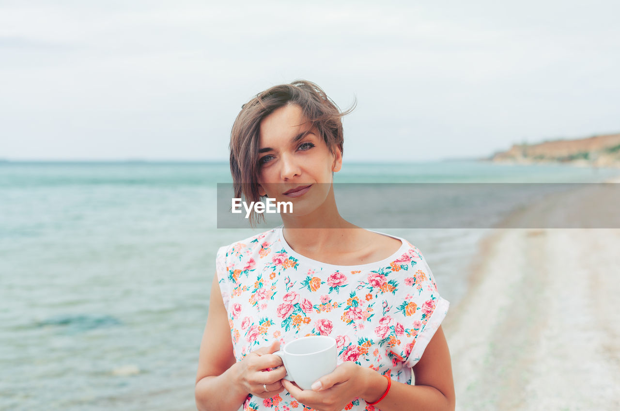 Portrait of woman holding coffee cup while standing against sea