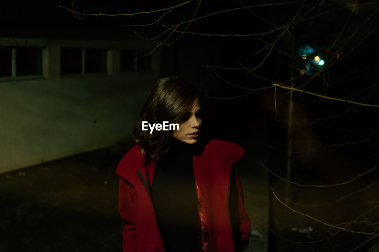 YOUNG WOMAN LOOKING AWAY WHILE STANDING AGAINST ILLUMINATED LIGHT