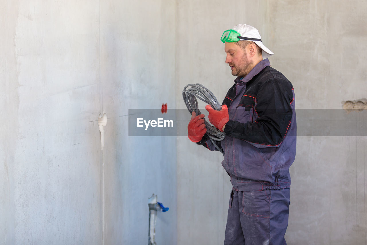 An electrician stands against the wall with an electrical cable for sockets.