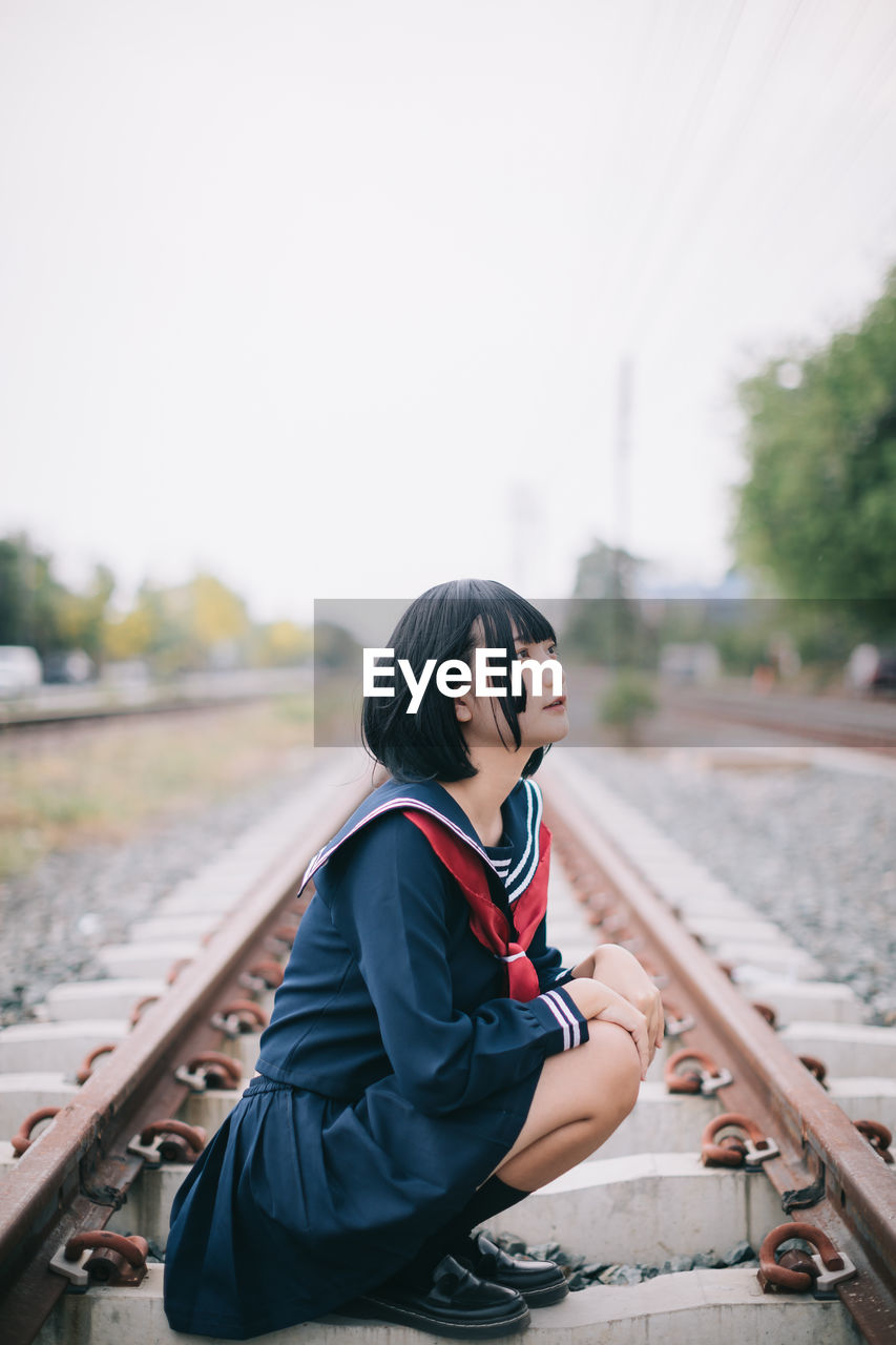 Woman looking away while sitting on railroad track against clear sky