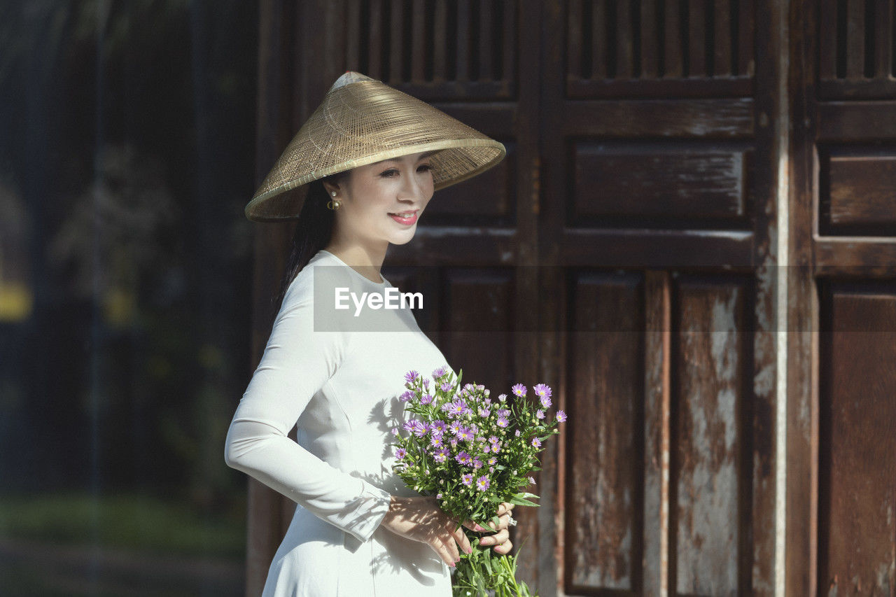women, flower, adult, flowering plant, one person, clothing, plant, young adult, fashion accessory, hat, nature, bouquet, smiling, standing, fashion, flower arrangement, happiness, dress, female, bride, emotion, portrait, beauty in nature, spring, celebration, wedding dress, lifestyles, three quarter length, sun hat, white, outdoors, architecture, looking, person, holding, elegance, wood, day, event, looking away, wedding, side view, freshness, waist up, positive emotion, fedora