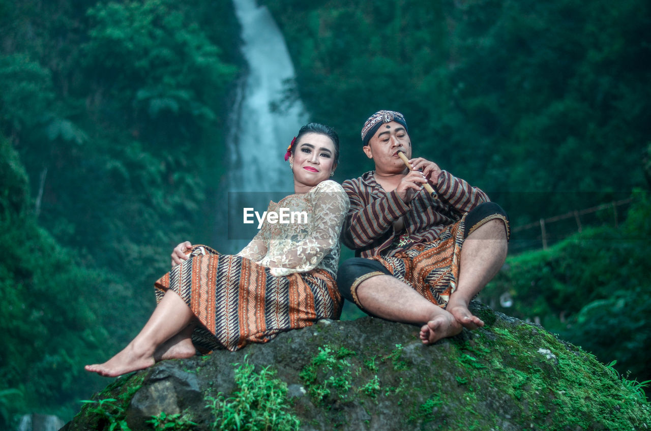 Smiling couple sitting on rock against waterfall