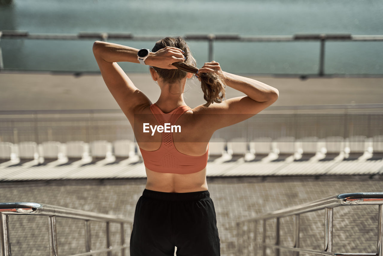 Back view of unrecognizable slim female athlete in sportswear standing putting on hairband on urban embankment near river and relaxing after exercising in summer day