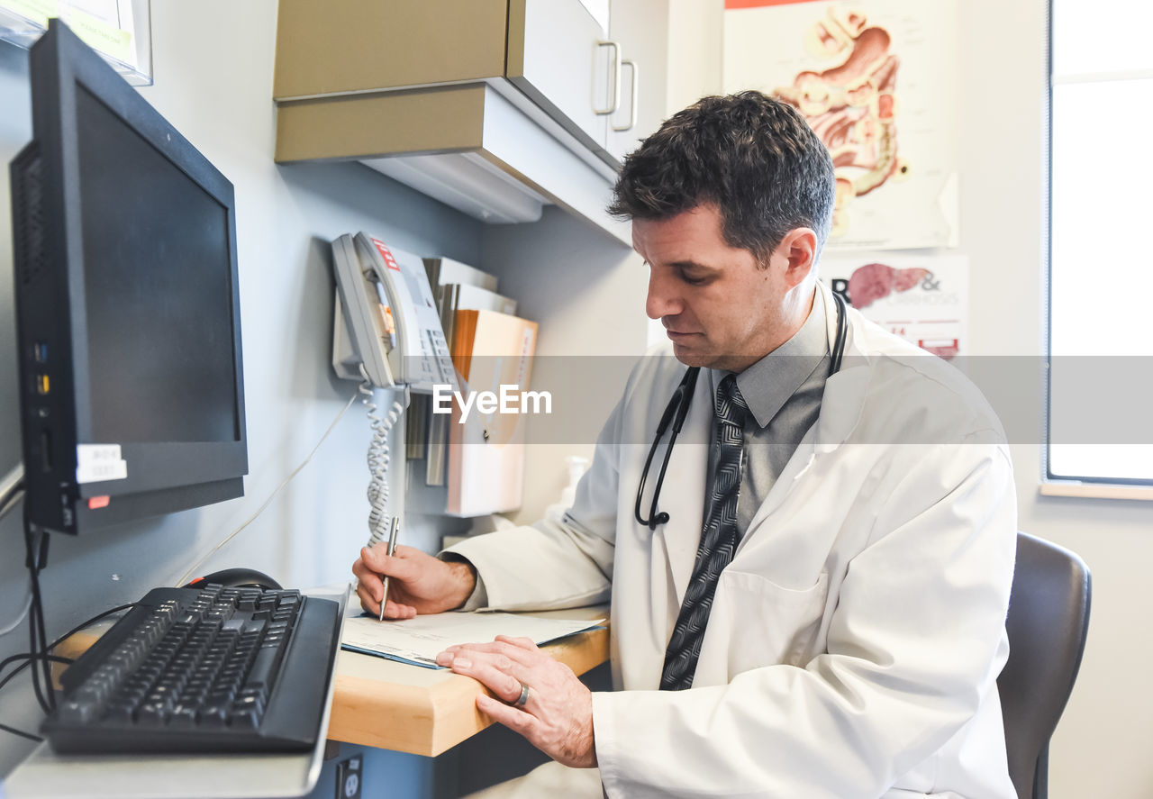 Doctor in a white coat writing in a file at a desk in a clinic room.