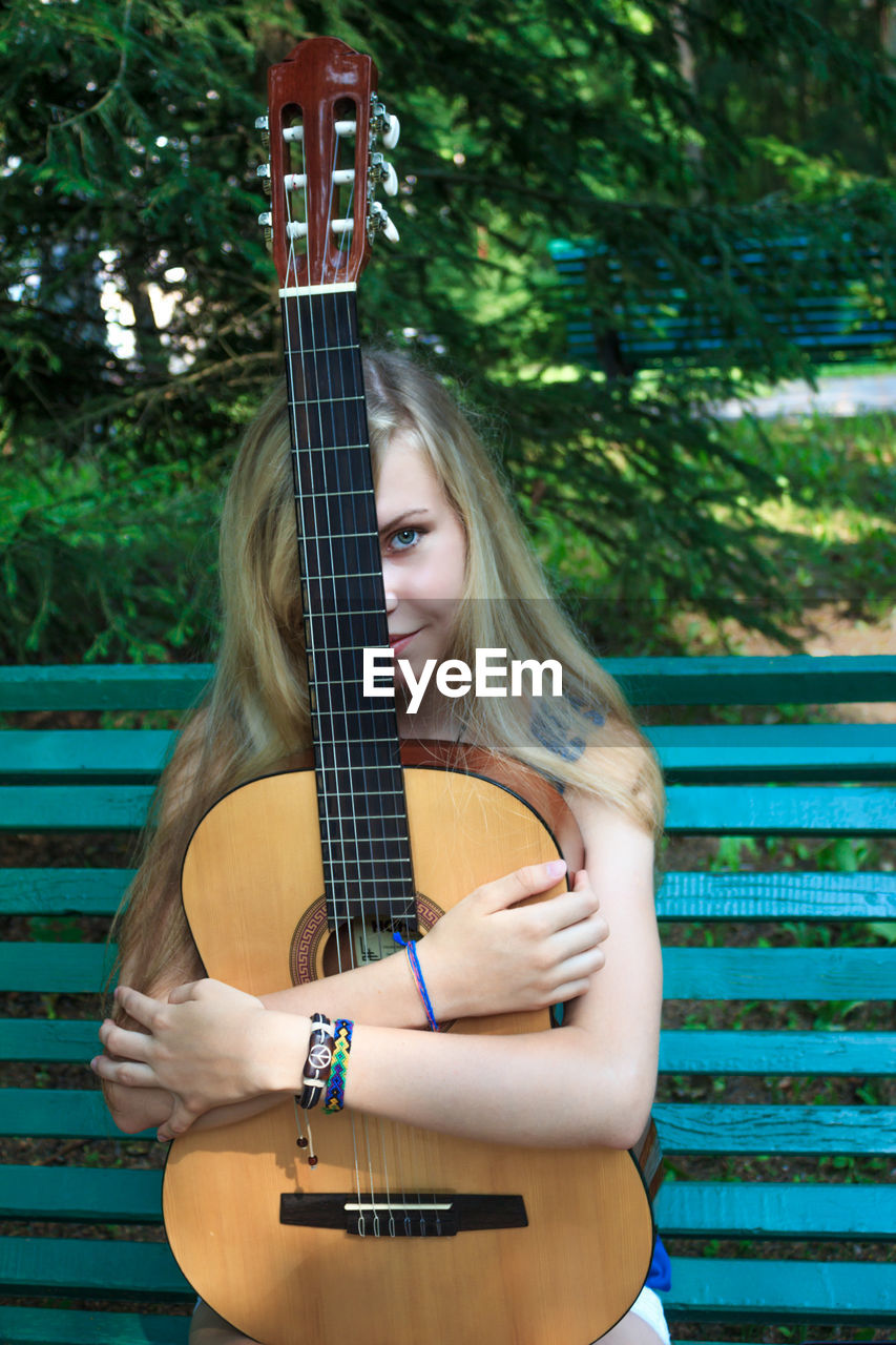 Portrait of young woman holding guitar while sitting in bench at park