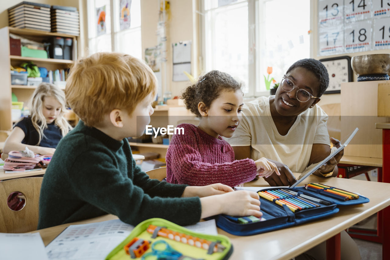 Smiling teacher assisting boy and girl with digital tablet sitting at desk in classroom