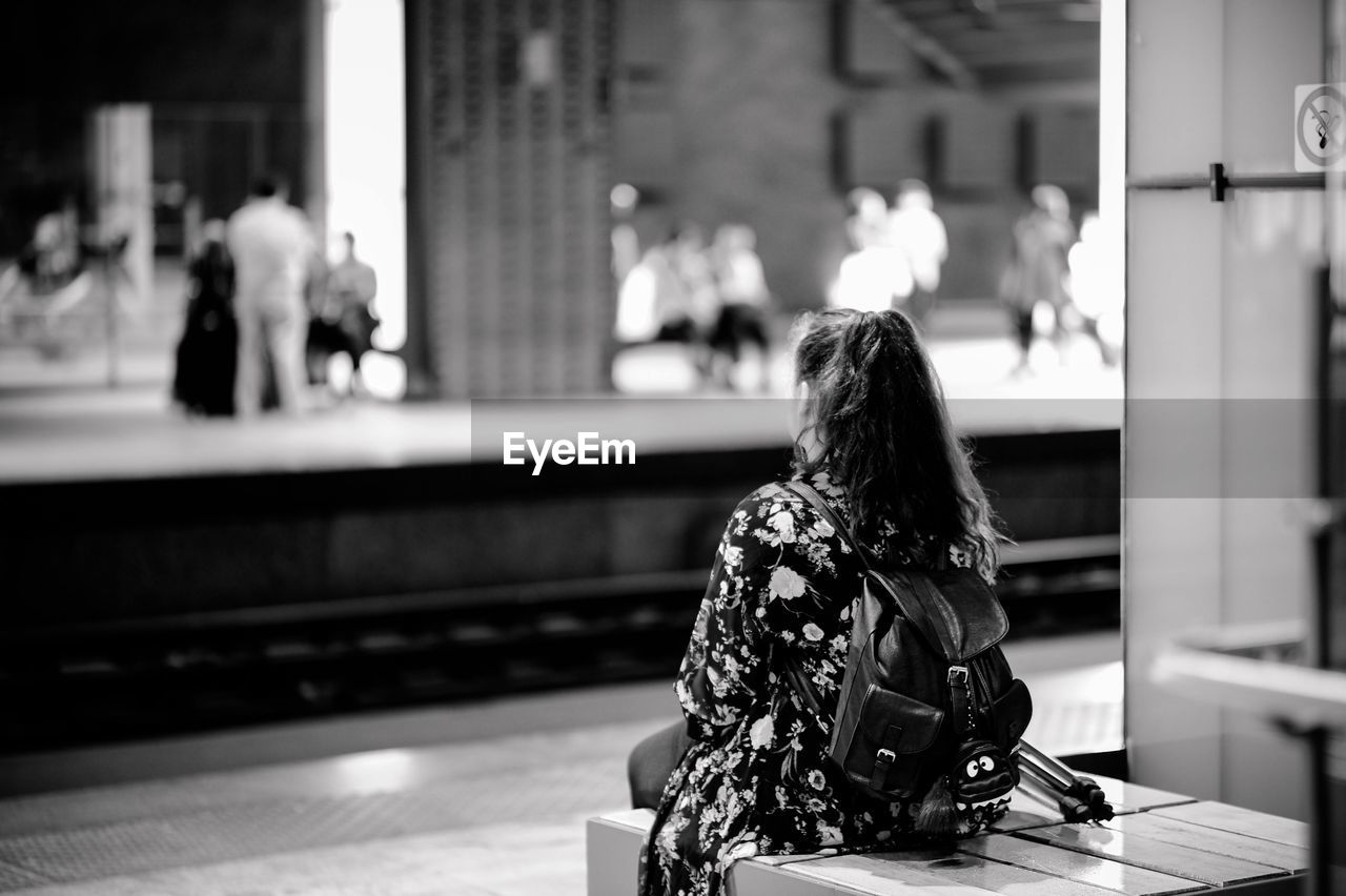 Rear view of young woman with backpack sitting at railroad station platform