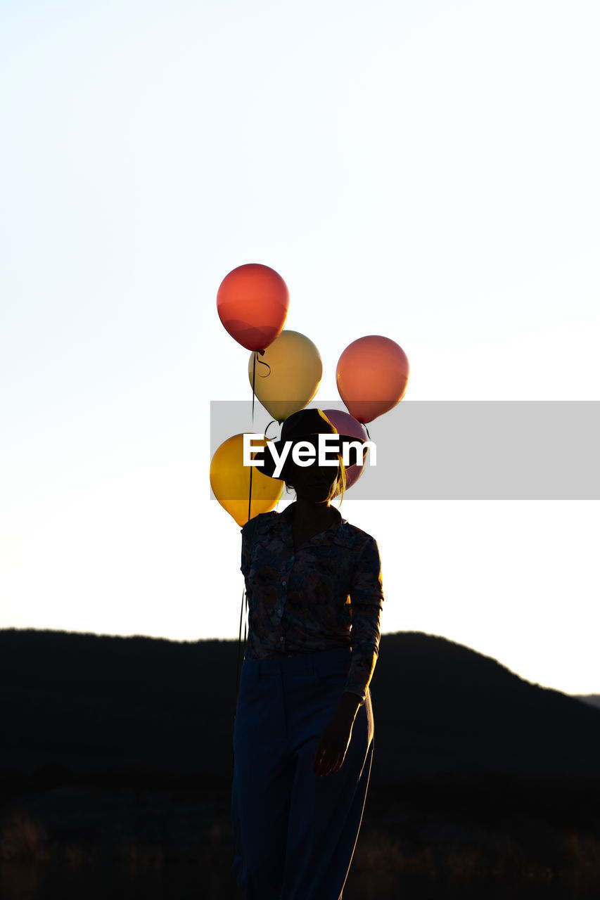 Silhouette of unrecognizable female in hat standing with heap of colorful balloons against cloudless sky at sunset time against mountain ridge