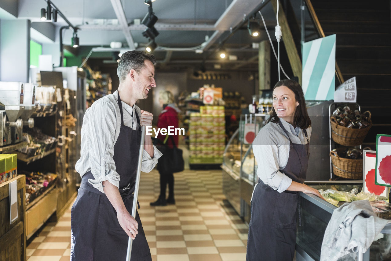 Smiling sales clerk holding broom while looking at saleswoman in supermarket