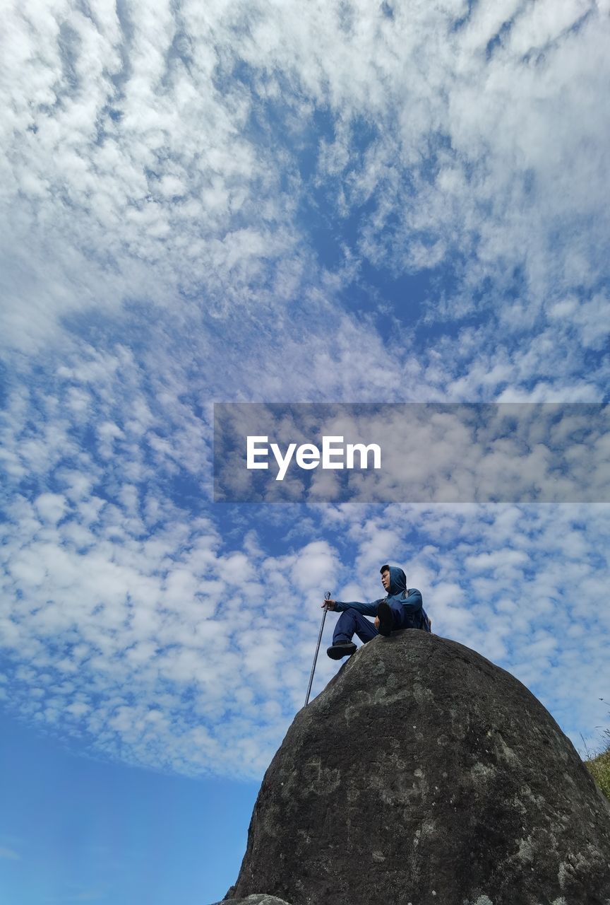 LOW ANGLE VIEW OF WOMAN SITTING ON ROCKS AGAINST SKY