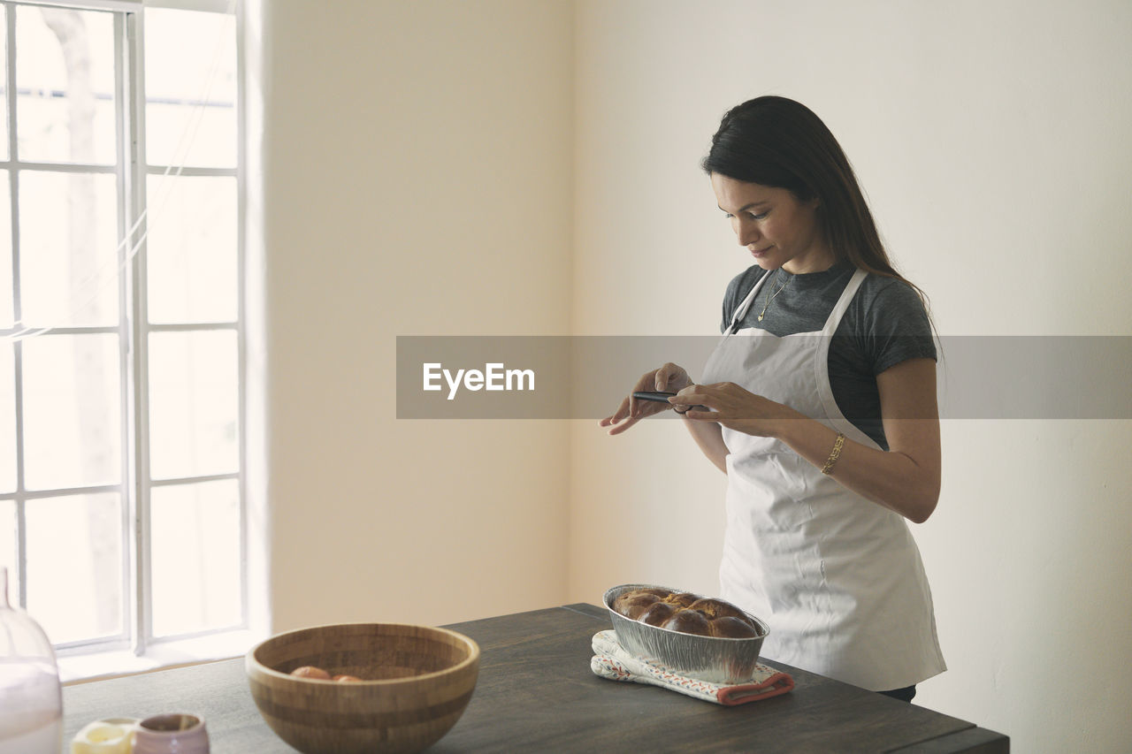 Woman in apron photographing baked challah bread with mobile phone on table against wall at home