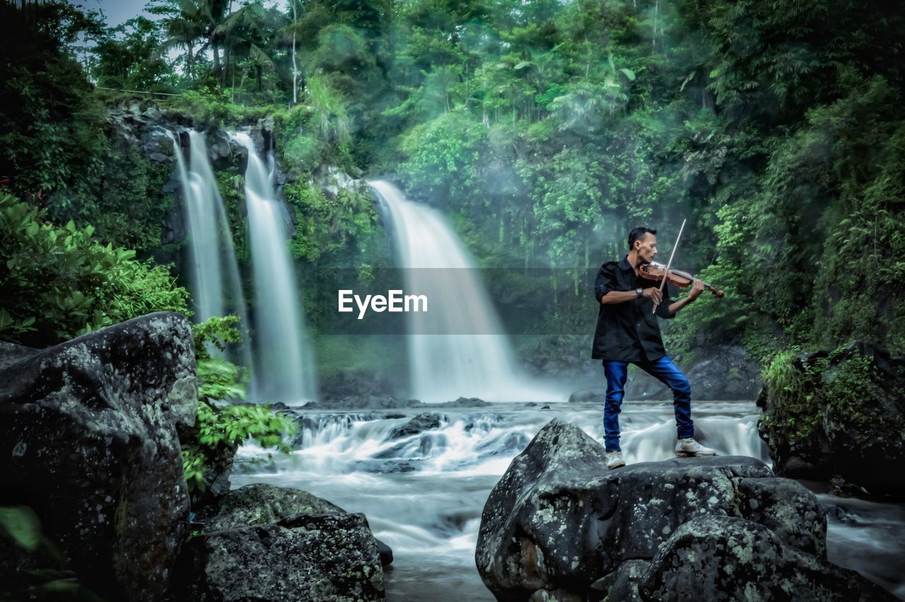 Man playing violin while standing on rock against waterfall in forest