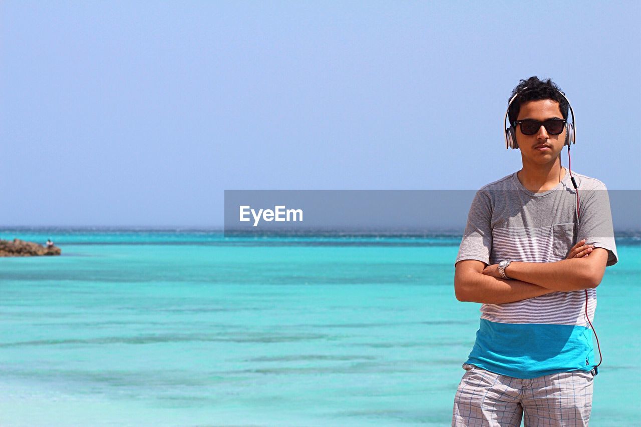 Portrait of young man listening to music while standing at beach against sky