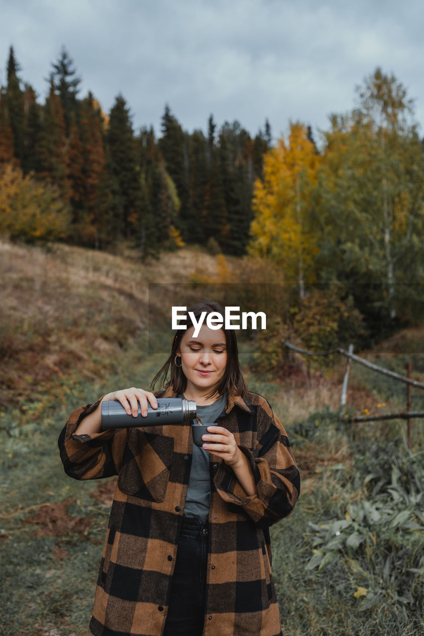 Portrait of smiling young woman standing on land with tea in thermos